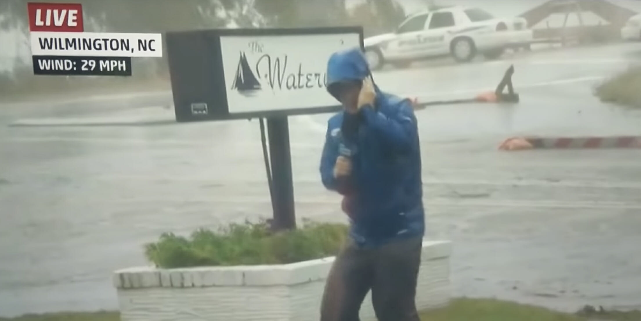 Weatherman dramatically braces against Hurricane Florence winds as dudes stroll casually in the background.