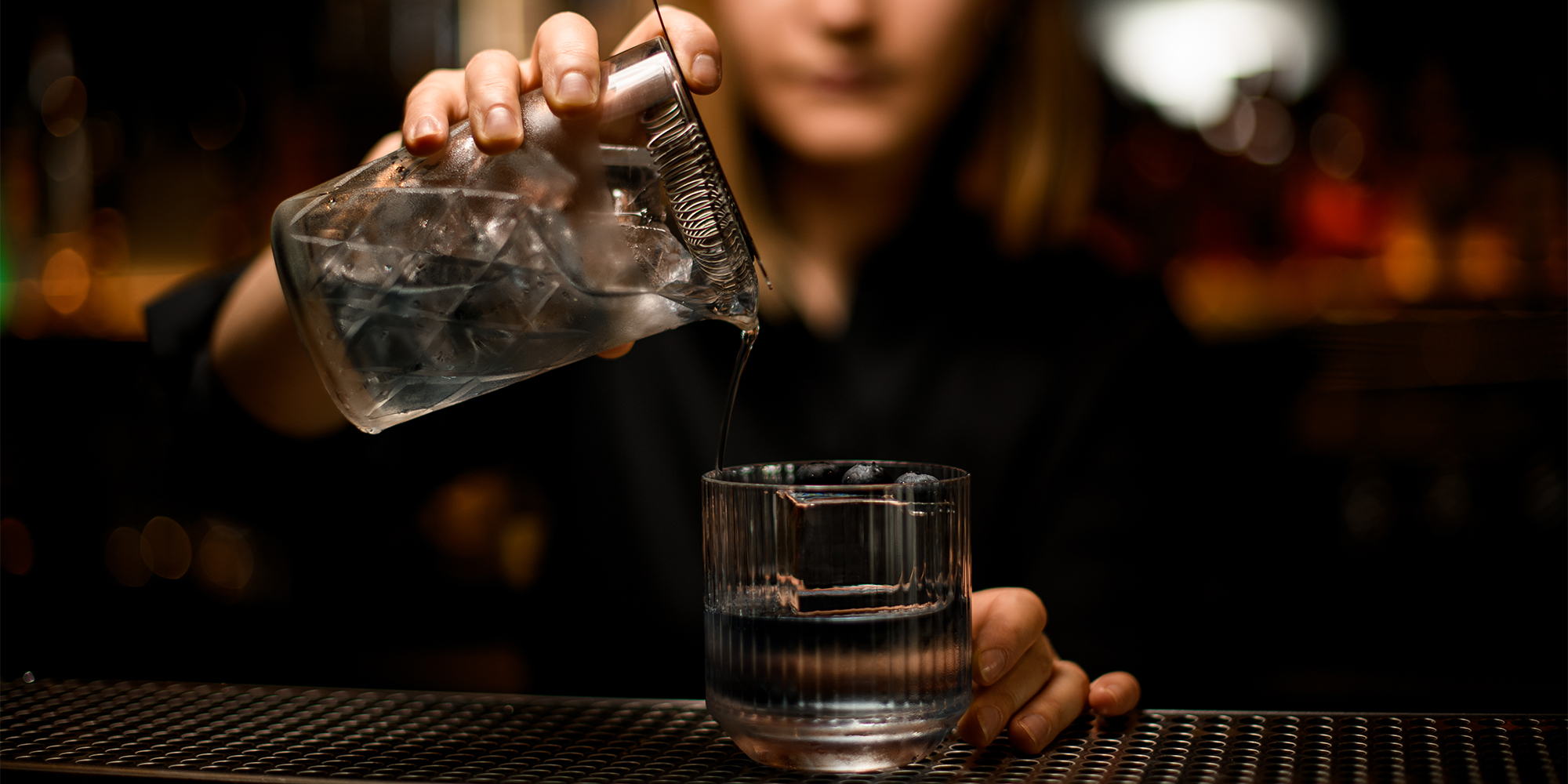 woman bartender holds mixing cup with cocktail and pours it into old-fashioned transparent glass with ice cube and blueberries
