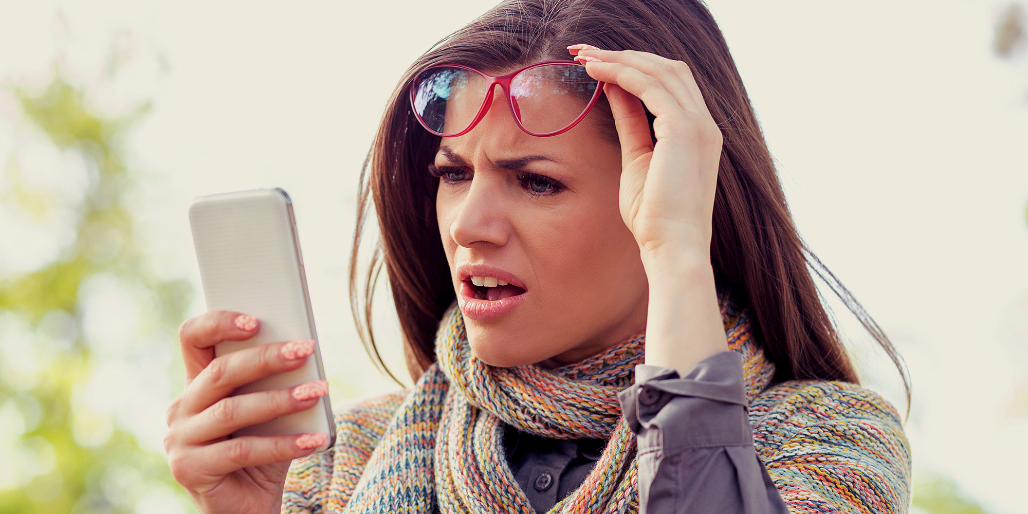 Annoyed upset woman in glasses looking at her smart phone with frustration while walking on a street on an autumn day