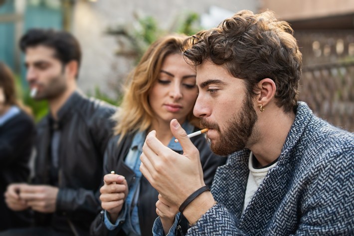 Group of friends lighting cigarettes and smoking together sitting on a bench outdoors