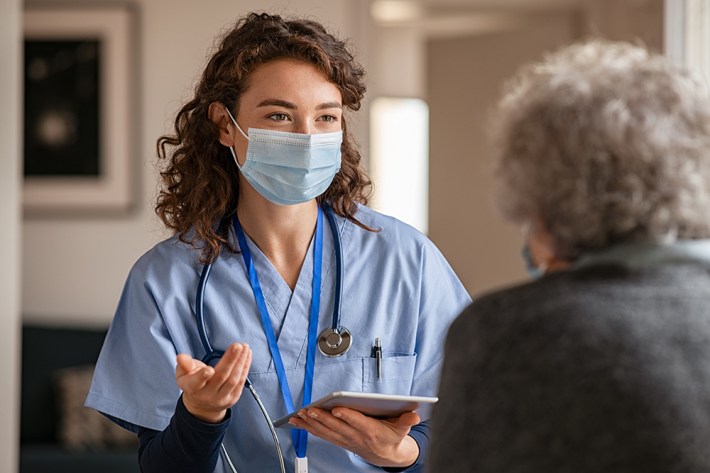Doctor wearing safety protective mask supporting and cheering up senior patient during home visit during covid-19 pandemic. Nurse and old woman wearing facemasks during coronavirus and flu outbreak.