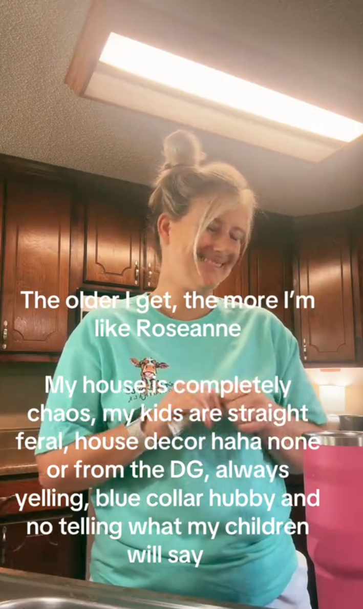 Screenshot of a blonde woman smiling in her kitchen as she prepares a drink. Text overlay reads, "The older I get, the more I'm like Roseanne. My house is completely chaos, my kids are straight feral, house decor haha none or from the DG, always yelling, blue collar hubby and no telling what my children will say."