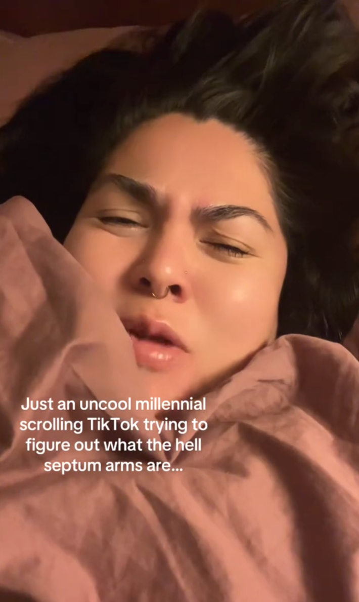 A woman cozy in a pink blanket looking confused. Text overlay reads, "Just an uncool millennial scrolling TikTok trying to figure out what the hell septum arms are..."