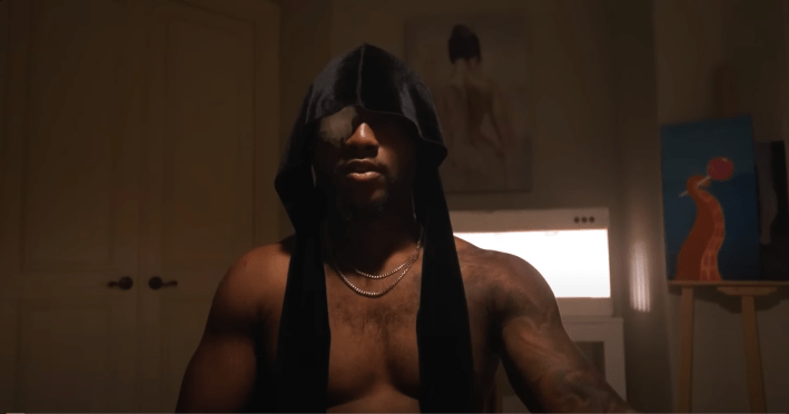 Screenshot of a video of shirtless streamer YourRAGE wearing an untied durag on his head, a bandage covering one eye. There is dramatic overhead lighting.