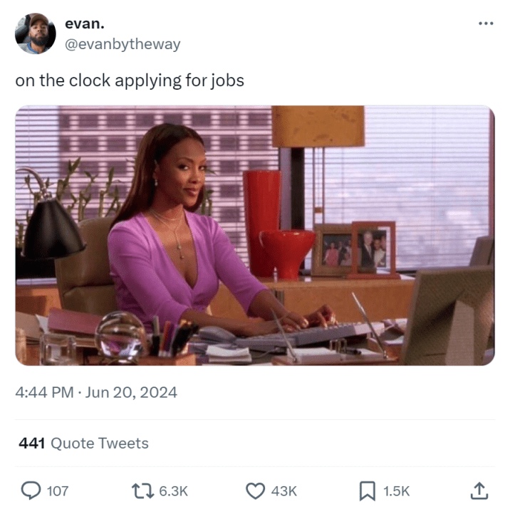 Photo of a woman smirking while sitting at her desk. Text reads, "on the clock applying for jobs."