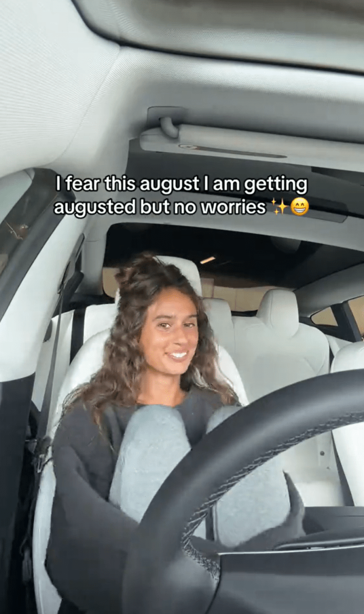 A woman sad but smiling for the camera sitting in her car with her knees up . Text overlay reads, "I fear this august I am getting augusted but no worries"