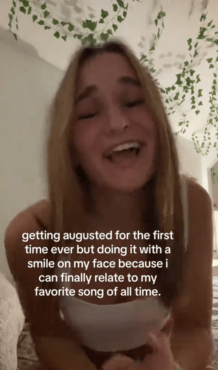 A blonde woman sitting in her bedroom singing for the camera and smiling. Text overlay reads, "getting augusted for the first time ever but doing it with a smile on my face because i can finally relate to my favorite song of all time."