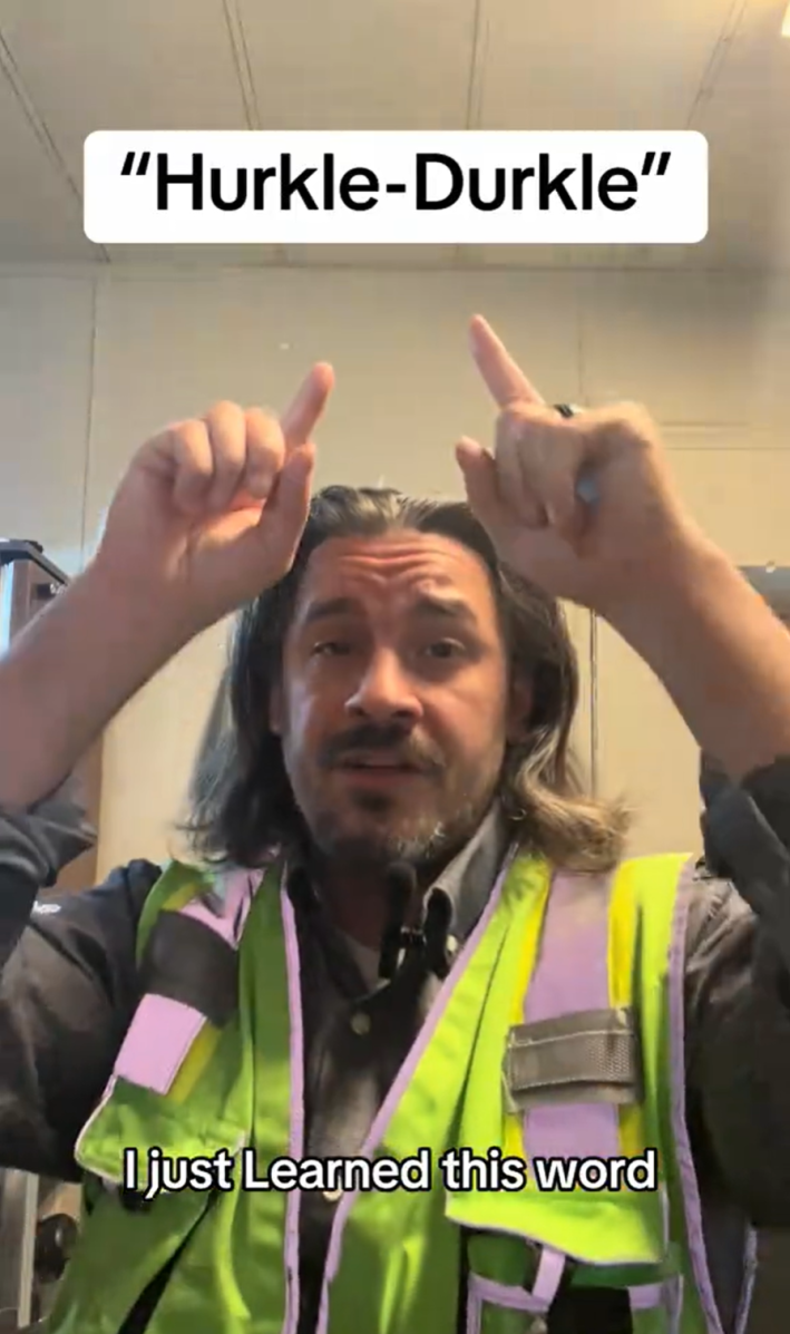 A man with long hair in a hi-viz vest pointing at the words"hurkle-durkle" on the screen.