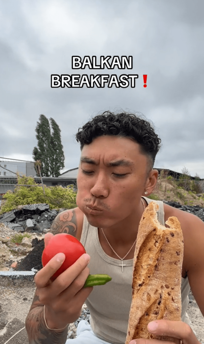 A man sitting on the ground outside, a baguette in one hand and a raw tomato and green pepper in the other. Text overlay reads, "Balkan Breakfast!"