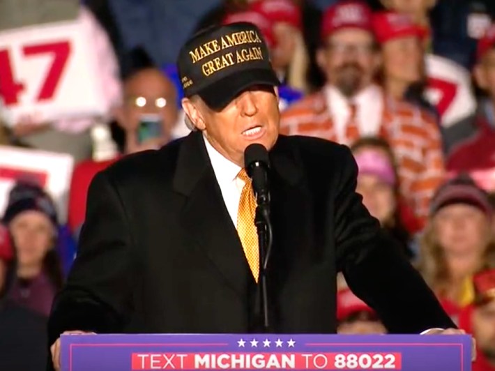 Donald Trump speaking in front of a Michiigan crowd. He stands at lectern and wears a new yellow and black "Make America Great AGain" hat.