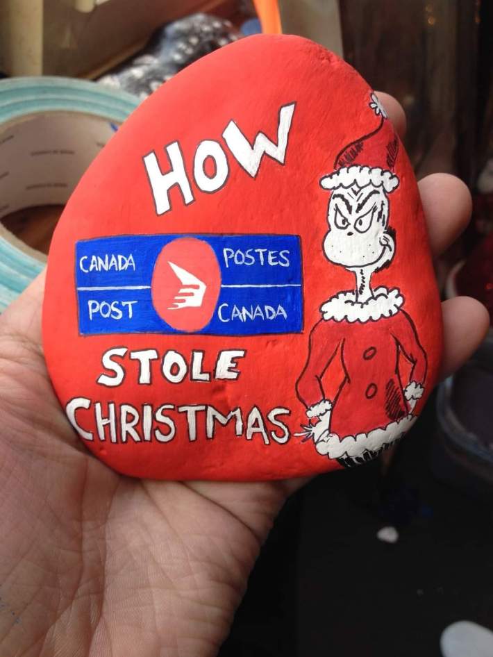 A painted rock in the Grinch's book art style, a drawing of the Grinch and Canada Post's logo. Text reads, "How Canada Post stole Christmas."