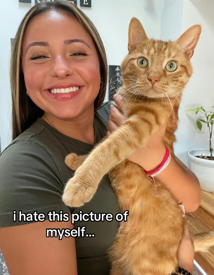TikTok photo of a young woman smiling for the camera holding an orange cat. Text overlay reads, "I hate this picture of myself..."