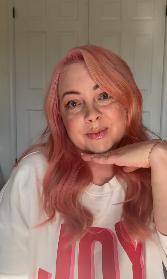 Makeup artist with pink hair in a "joy" shirt with one hand under her chin and smiling sadly for the camera.