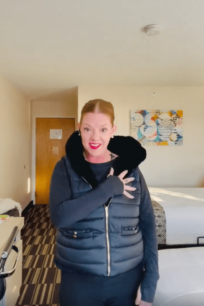 A woman in a puffer jacket smiling for the camera with her hand to her heart.