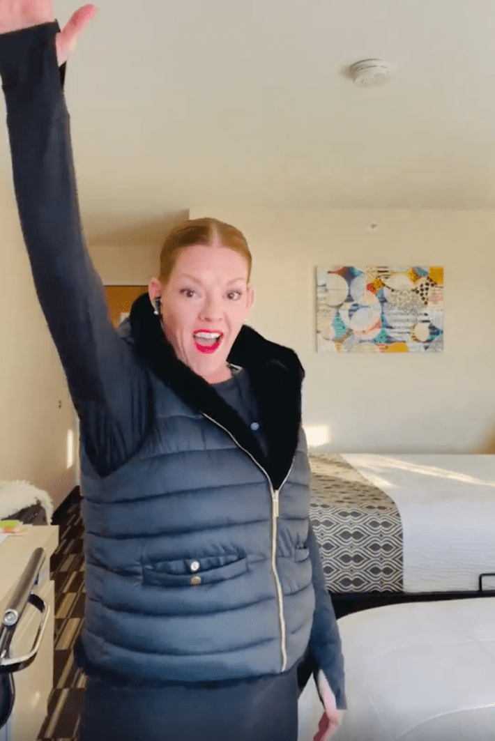 A woman in a puffer jacket smiling for the camera with her hand raised in a salute.