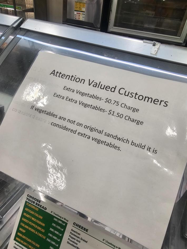Laminated printout that reads, "Attention Valued Customers: Extra Vegetables- $0.75 Charge. Extra Extra Vegetables-$1.50 Charge. If vegetables are not on original sandwich build it is considered extra vegetables."