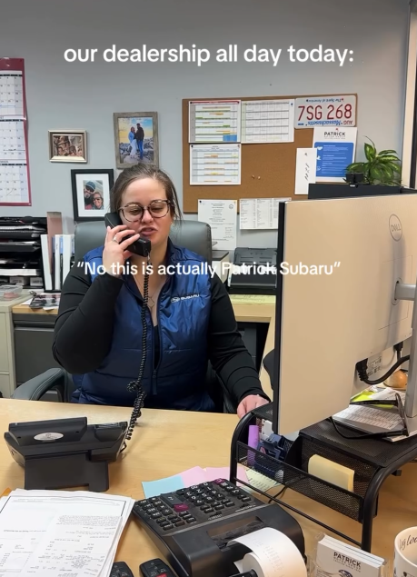 A woman in a vest sitting at the desk and speaking on a wired phone.