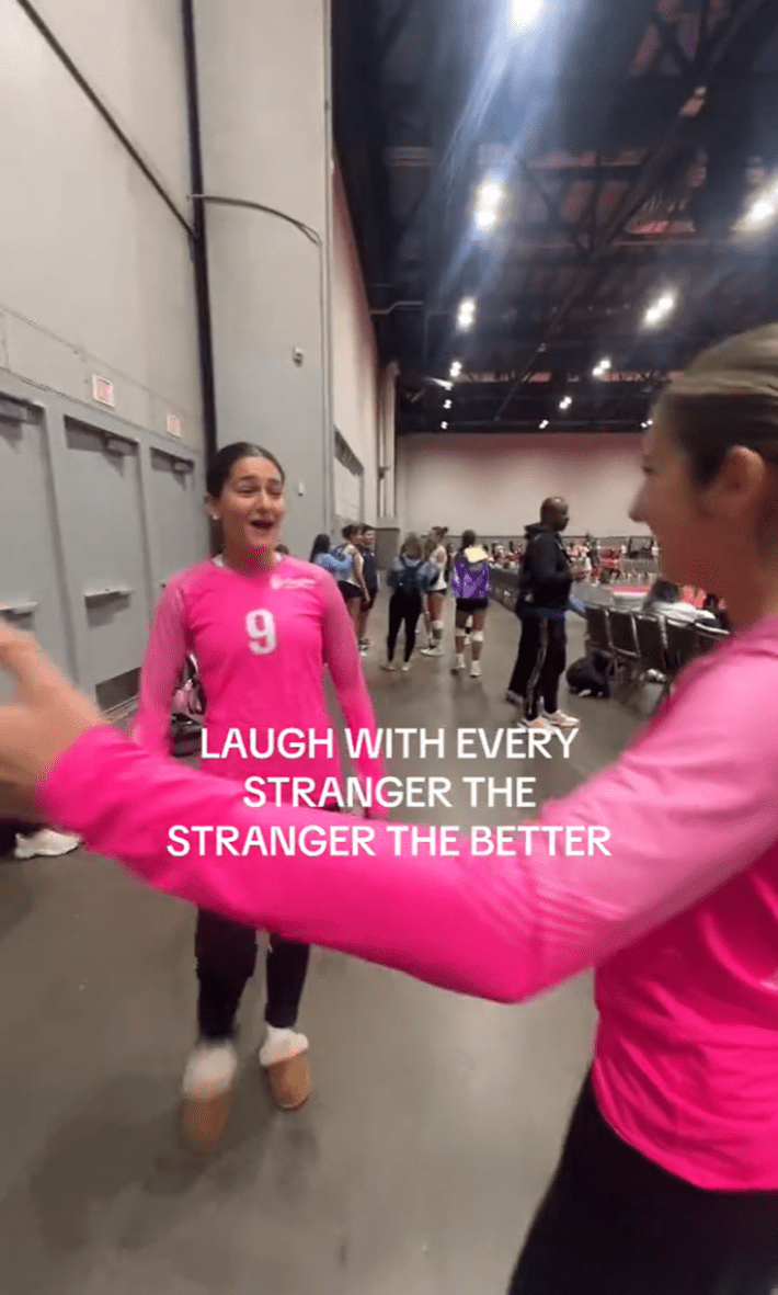 Laugh with every stranger trend, a girl in a pink fitted sports shirt opening her arms to hug a girl in a matching shirt at an event.
