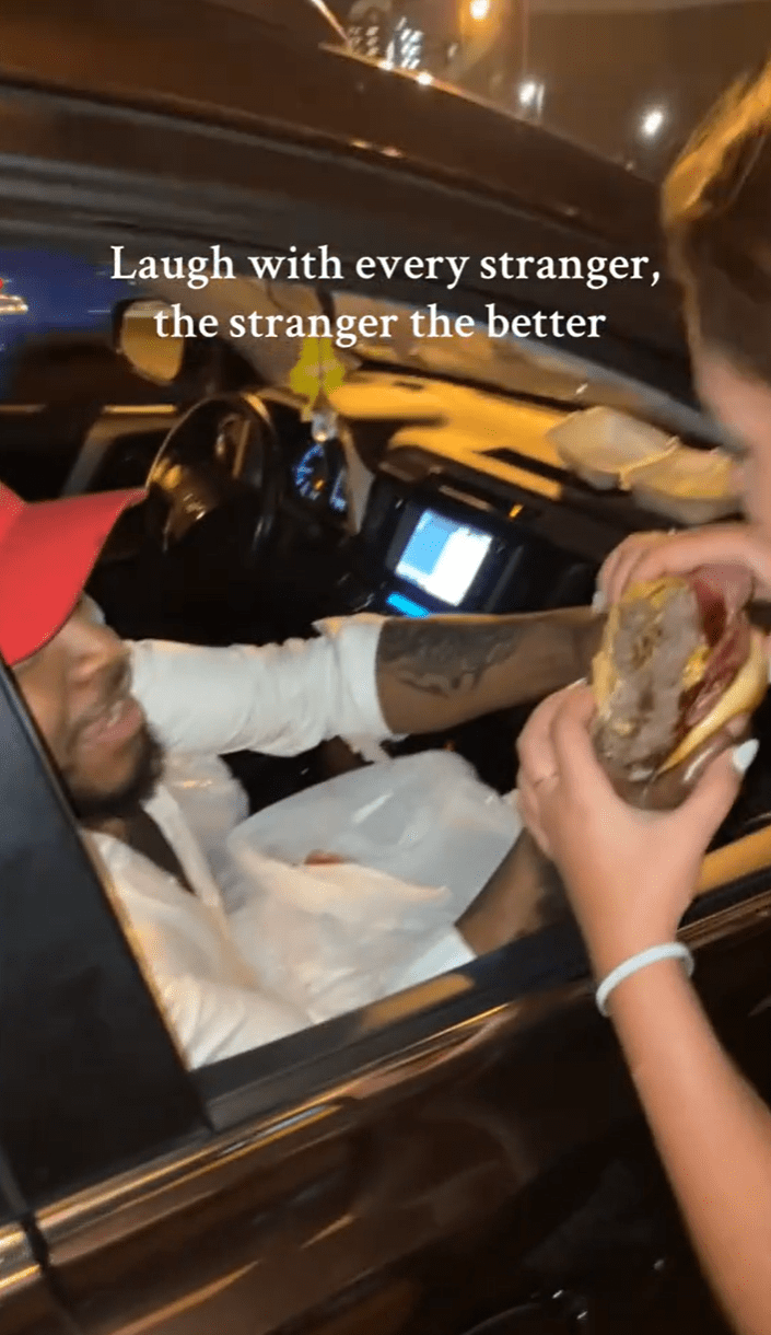 Laugh with every stranger trend, a woman eating a burger that a guy hands her from the passenger side of a car.