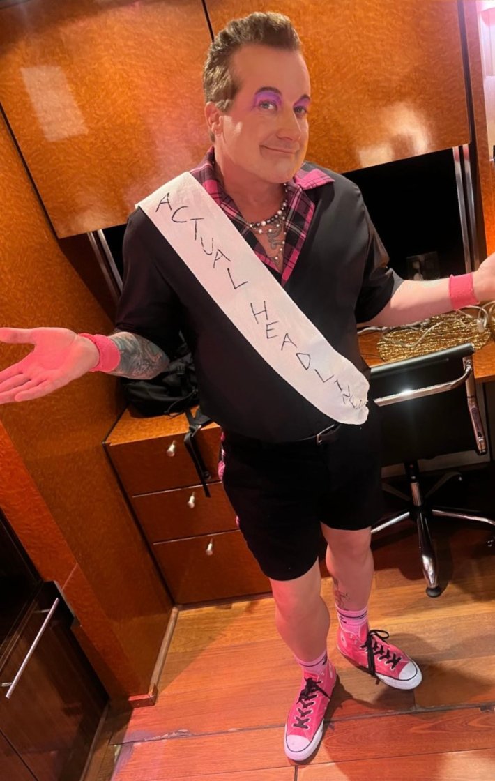 Tre Cool in black and pink punk outfit and flashy makeup wearing a sash that reads, "Actual Headliner" while shrugging his shoulders at the camera.