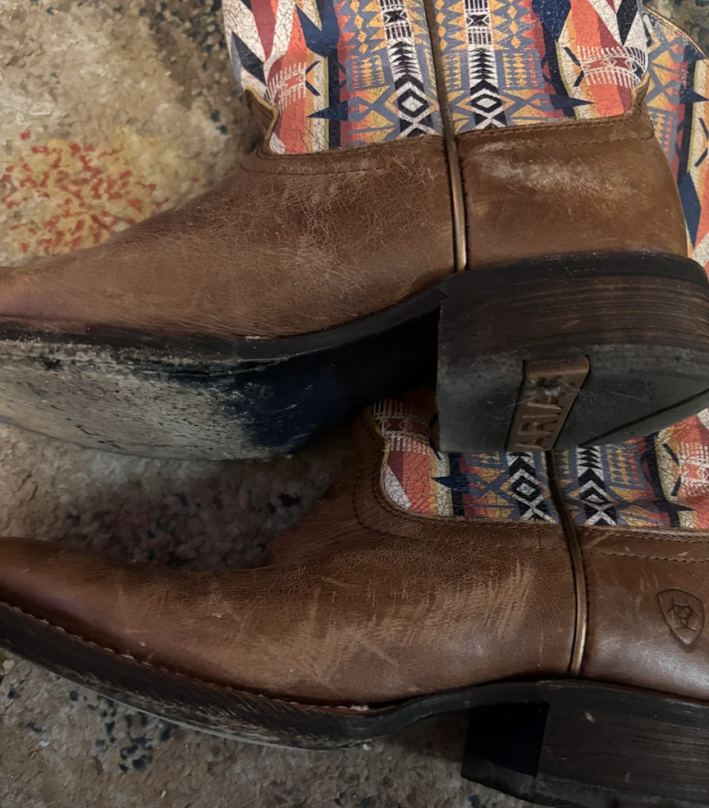 A photo of a pair of brown leather Ariat boots with patterned sides. The leather is cracked and scuffed and the soles are heavily worn out.