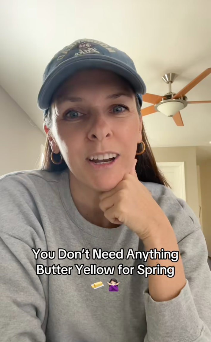 Woman in a grey crewneck and a baseball cap, chin resting on her closed fist, saying, "You don't need anything butter yellow for spring."