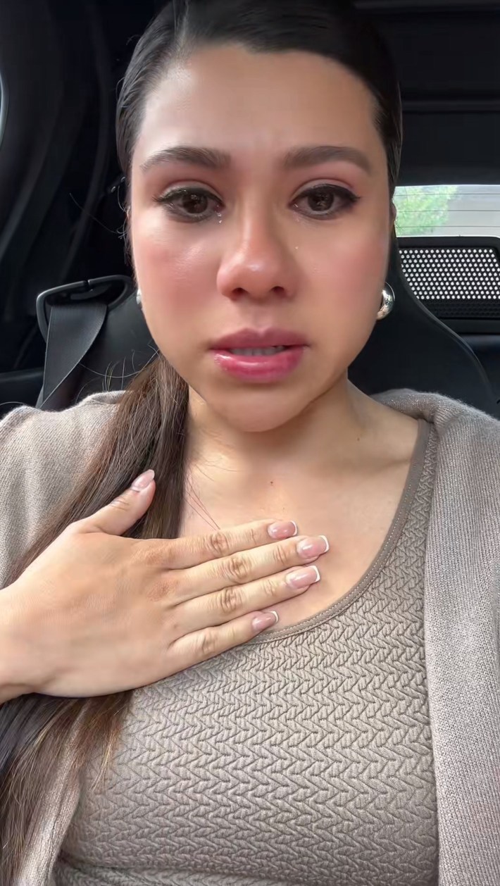 Tearful woman sitting in a car speaking to her phone, her hand on her chest.