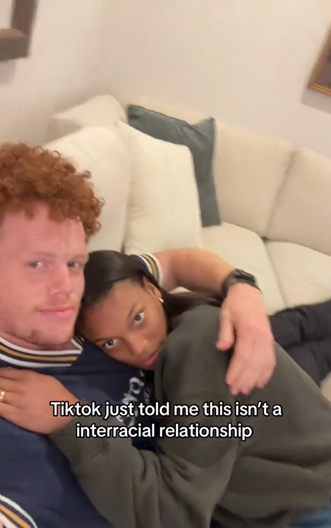 Redheaded man and his Black girlfriend cuddling on the couch. Text overlay reads, "TIkTok just told me this isn't a interracial relationship."