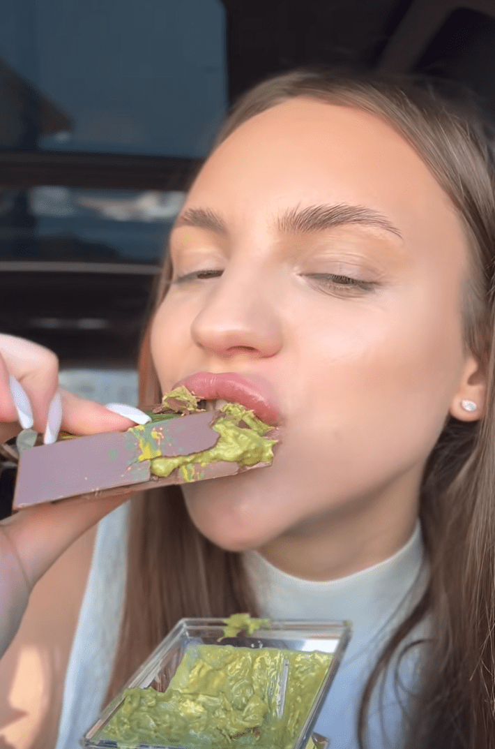 A close up of a woman eating a chocolate and pistachio bar that is falling apart.