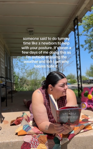 A woman lying outside with a book in the sun on her stomach. Text overlay reads, "Someone said to do tummy time like a newborn to help with your posture. It's been a few days of me doing this as I'm outside enjoying the weather and tbh I get why babies hate it."