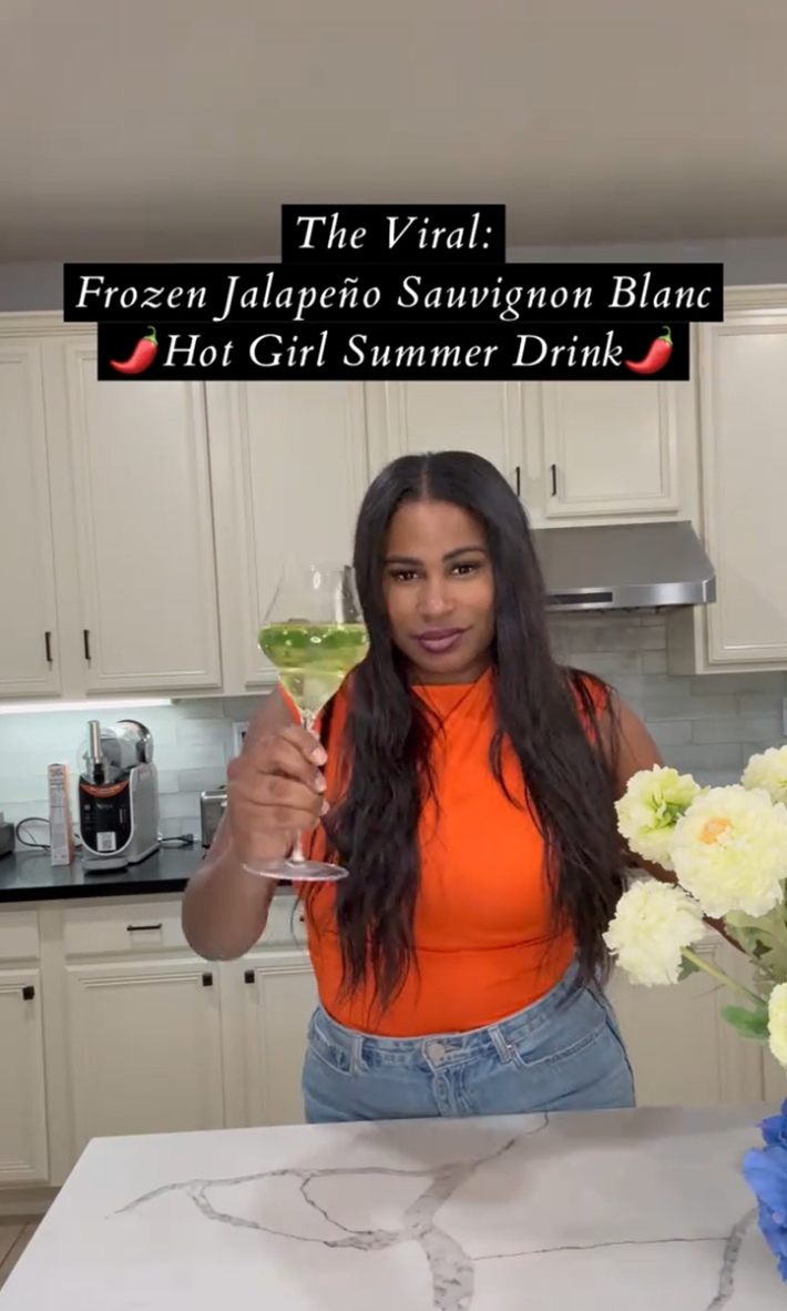 A woman standing in her kitchen cheersing the camera with her Spicy Sauvy B.