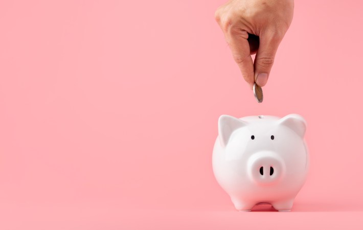 Money saving and investment concept. Hand putting coin to piggy bank on pink background with copy space