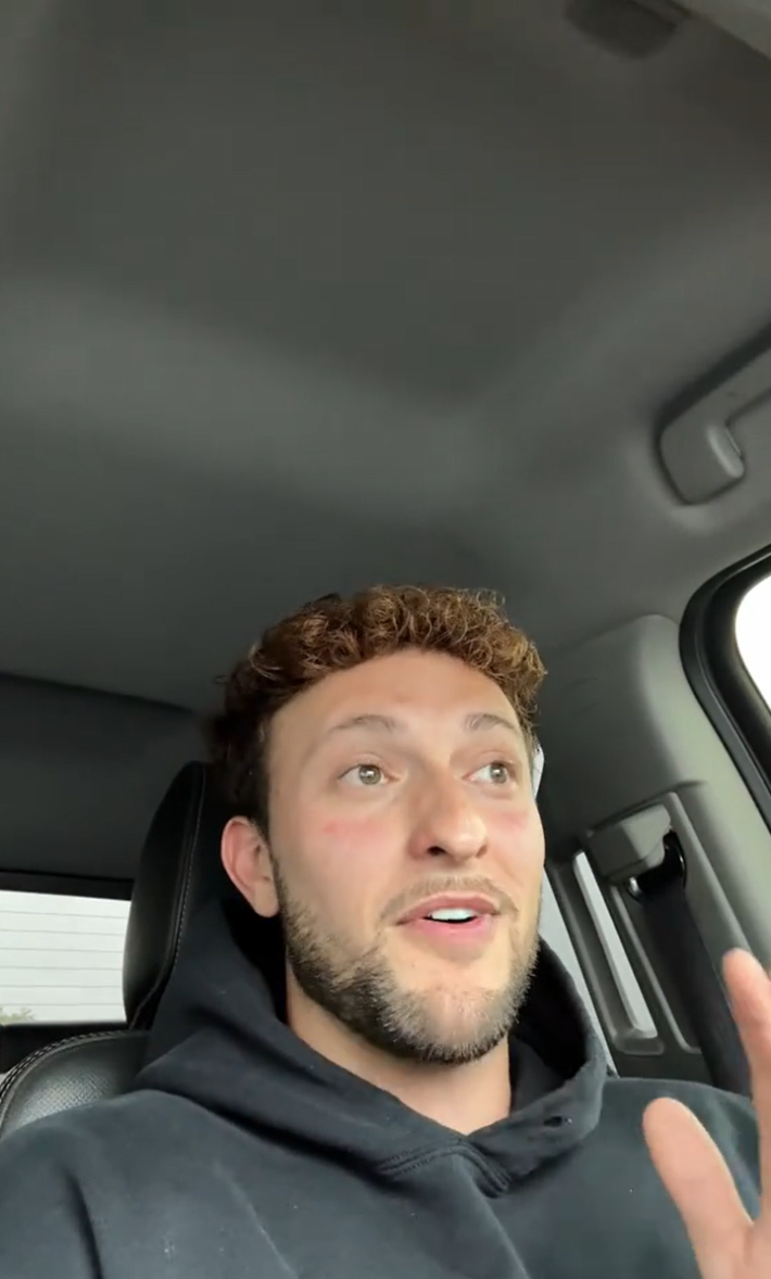 A man with curly red hair talking to the camera in his car, one hand raised.