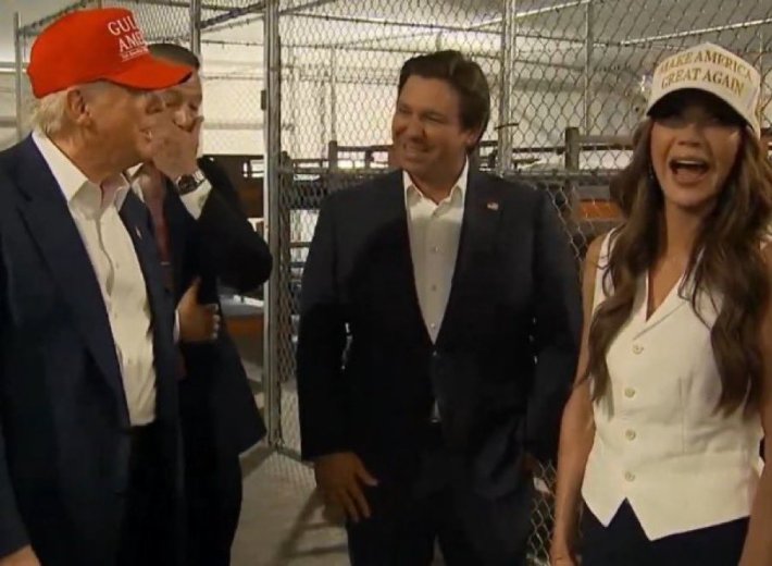 Slightly blurry photo of Trump and Republican leaders touring Alligator Alcatraz and laughing in front of cages that will house people they are deporting.
