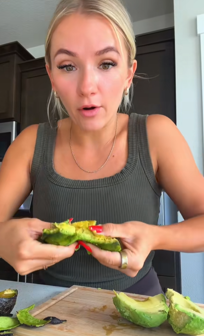 Blonde woman in the kitchen trying to break a Costco avocado for the camera to prove how rubbery it is.