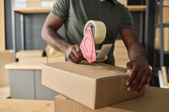 Close-up of man packing cardboard box with adhesive tape in storage room