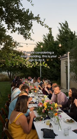 Intimate gathering of wedding guests all sat at a long table for the reception. Text overlay reads, "If you're considering a small wedding, this is your sign to do it."