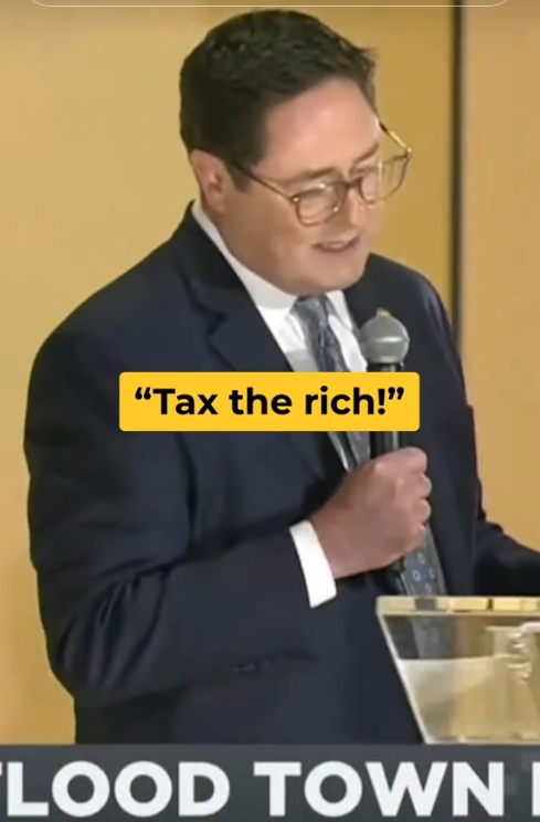 Rep. Mike Flood standing on stage with a mic and grimacing as the crowd chants "Tax the rich!"
