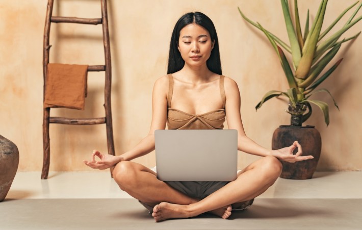 girl sitting in lotus pose with laptop, watching yoga tutorial online or using meditation app, following instructor, keeping hands in mudra sign