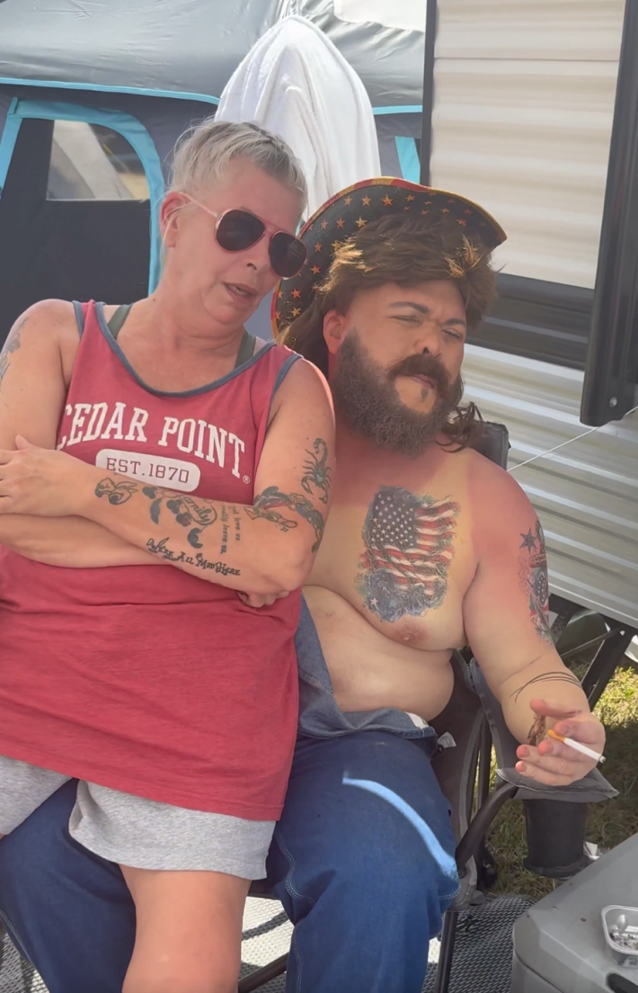 Druski with a white woman wearing a Cedar Point tank top sitting on his lap, sharing a cigarette.