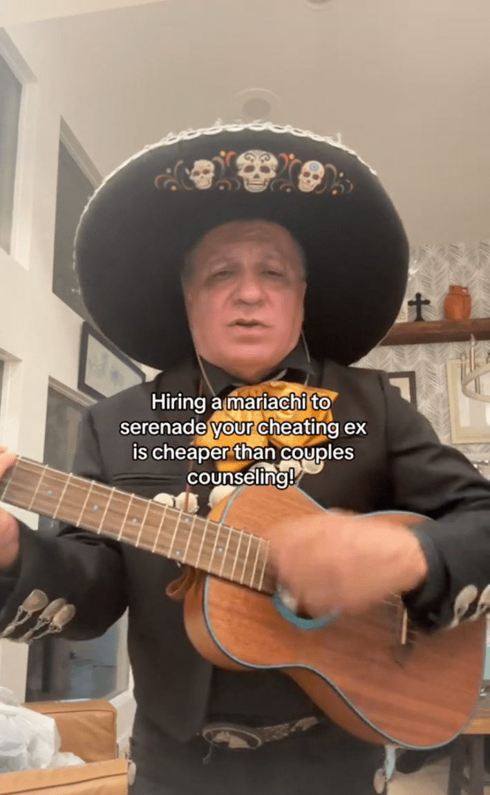 Mariachi performer strumming on a guitar, facing toward the camera. His hat has three skulls embroidered on it.