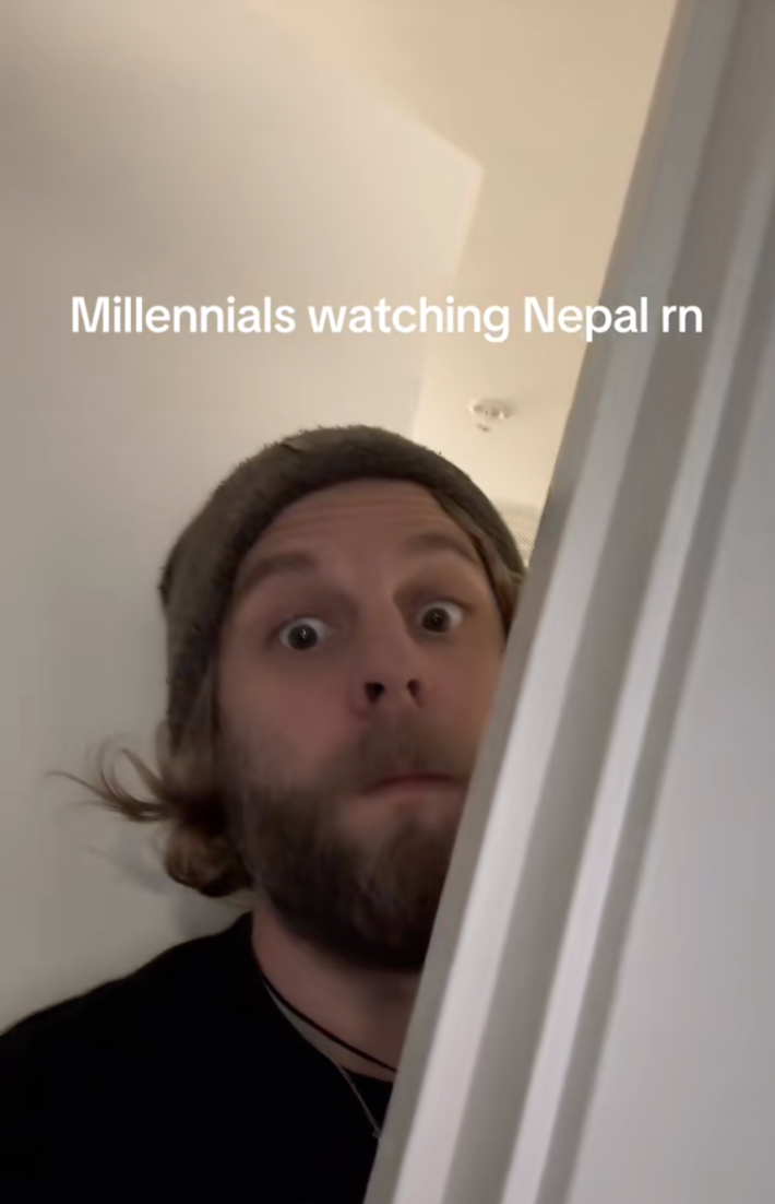 A man in a beanie staring around a doorway wide eyed. Text reads, "Millennials watching Nepal rn."