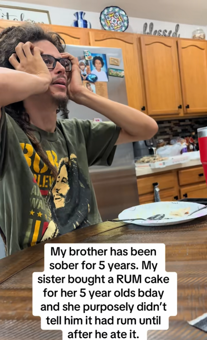 A man wearing a Bob Marley t-shirt clutches his head in the kitchen after learning he was fed rum cake by his sister.