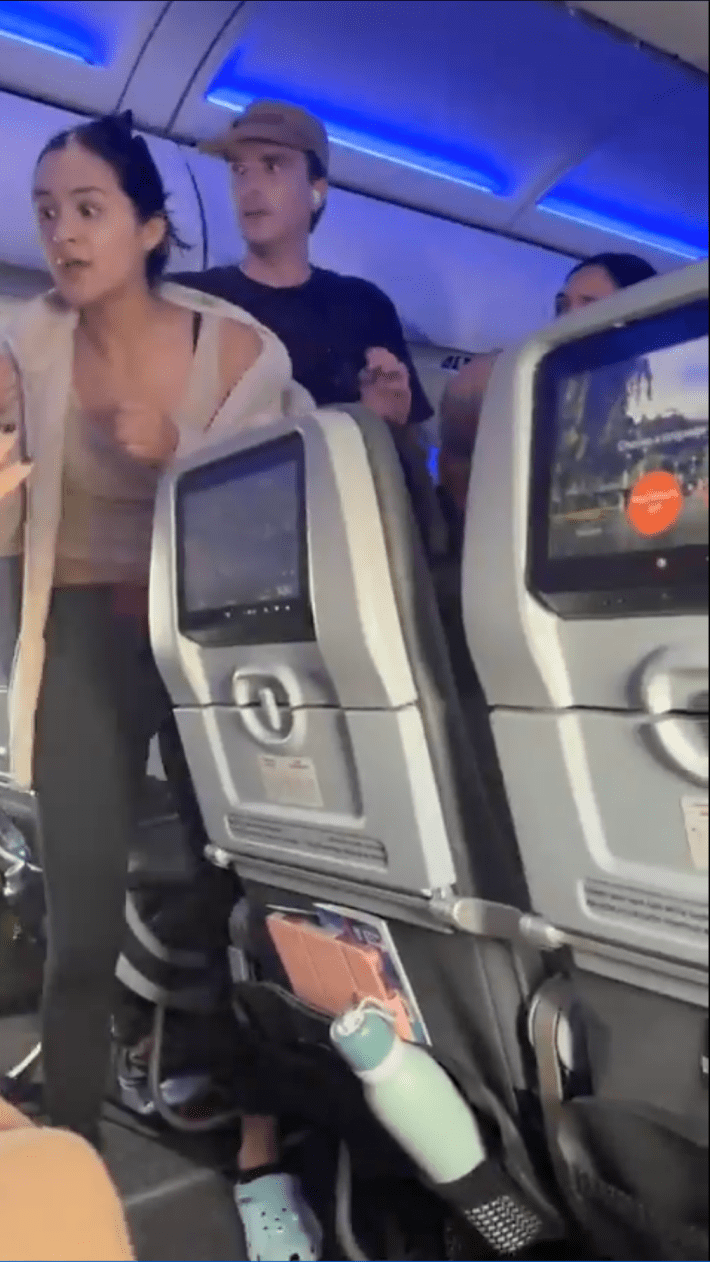 A woman looking visibly distressed in the airplane aisle while other passengers and fight attendants try to calm her.