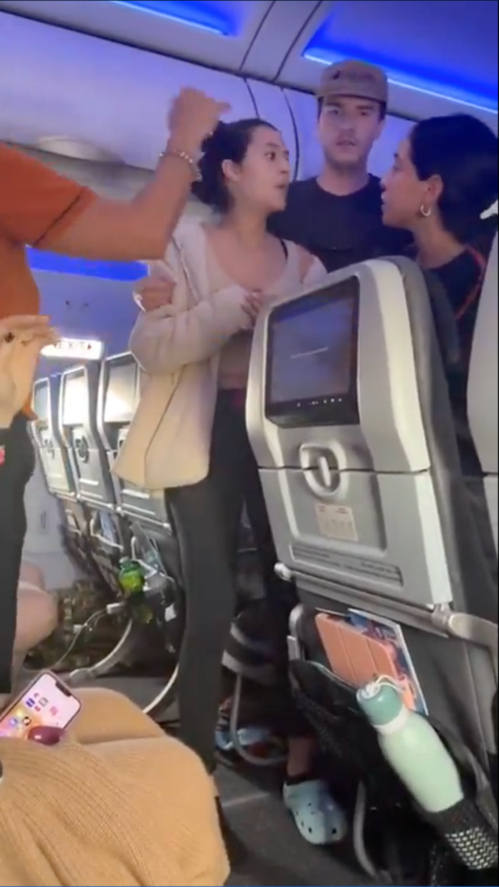 A woman looking visibly distressed in the airplane aisle while other passengers and fight attendants try to calm her.