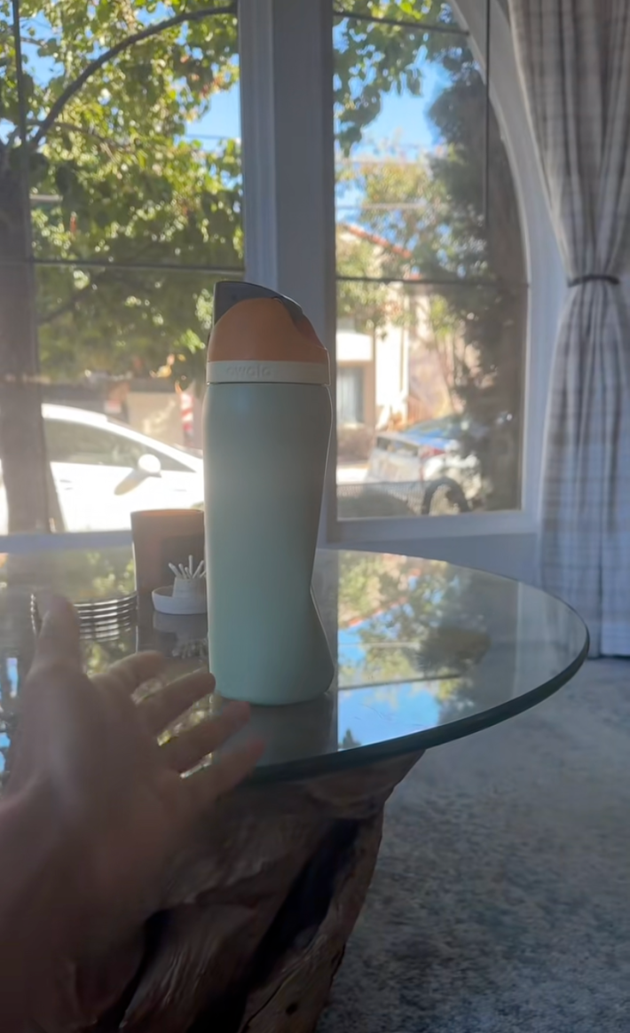 A dented Owala bottle sitting on a glass coffee table in a bright and sunny living room.