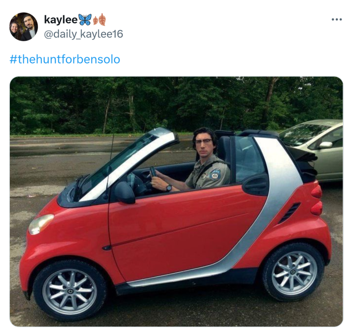 Adam Driver in a red Smart Car staring at the camera seriously.