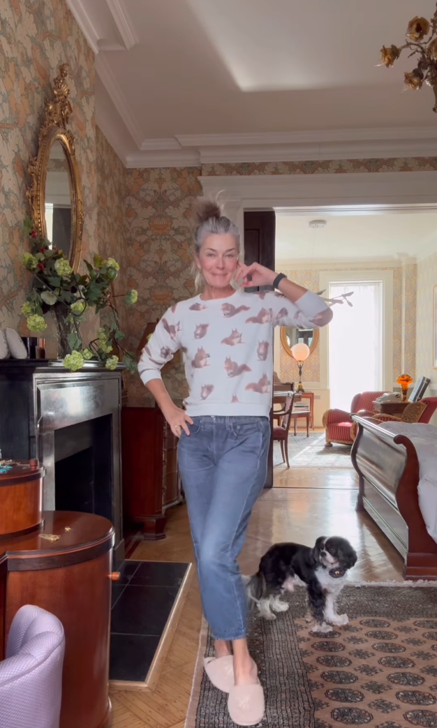 60 year old supermodel posing in her comfortable outfit of stretchy jeans and squirrel sweater, her dog looking on from behind her.