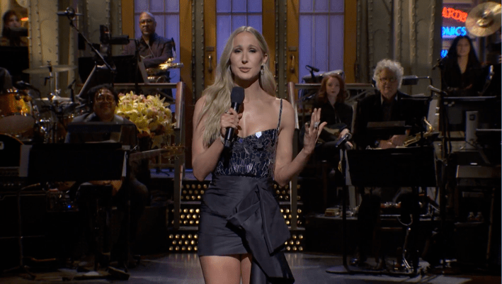 Nikki Glaser hosts SNL. She is standing on stage holding a mic as she does her monologue, with a range of people sitting behind her.