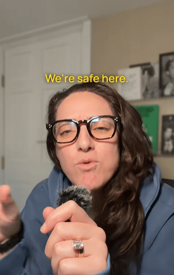 Woman with long brown hair and big glasses holding a lapel mic in her hand and talking to the camera in her home. Text overlay reads, "We're safe here."
